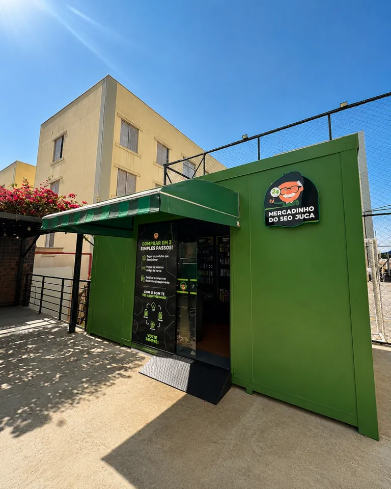 Fachada de uma loja externa do Seo Juca, verde, com toldo e porta de vidro, ao lado de um prédio