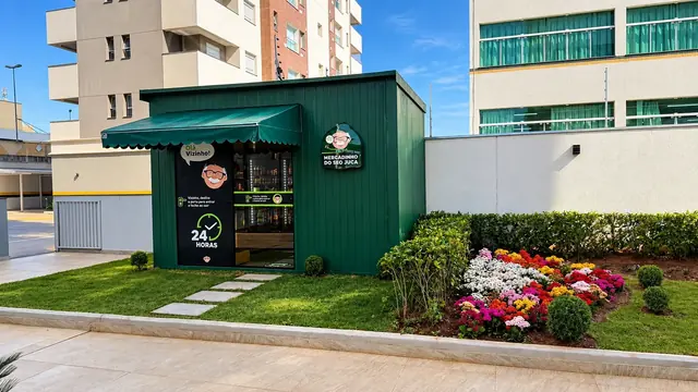 Loja modular do Seo Juca, verde, com toldo, jardim florido e prédios ao fundo